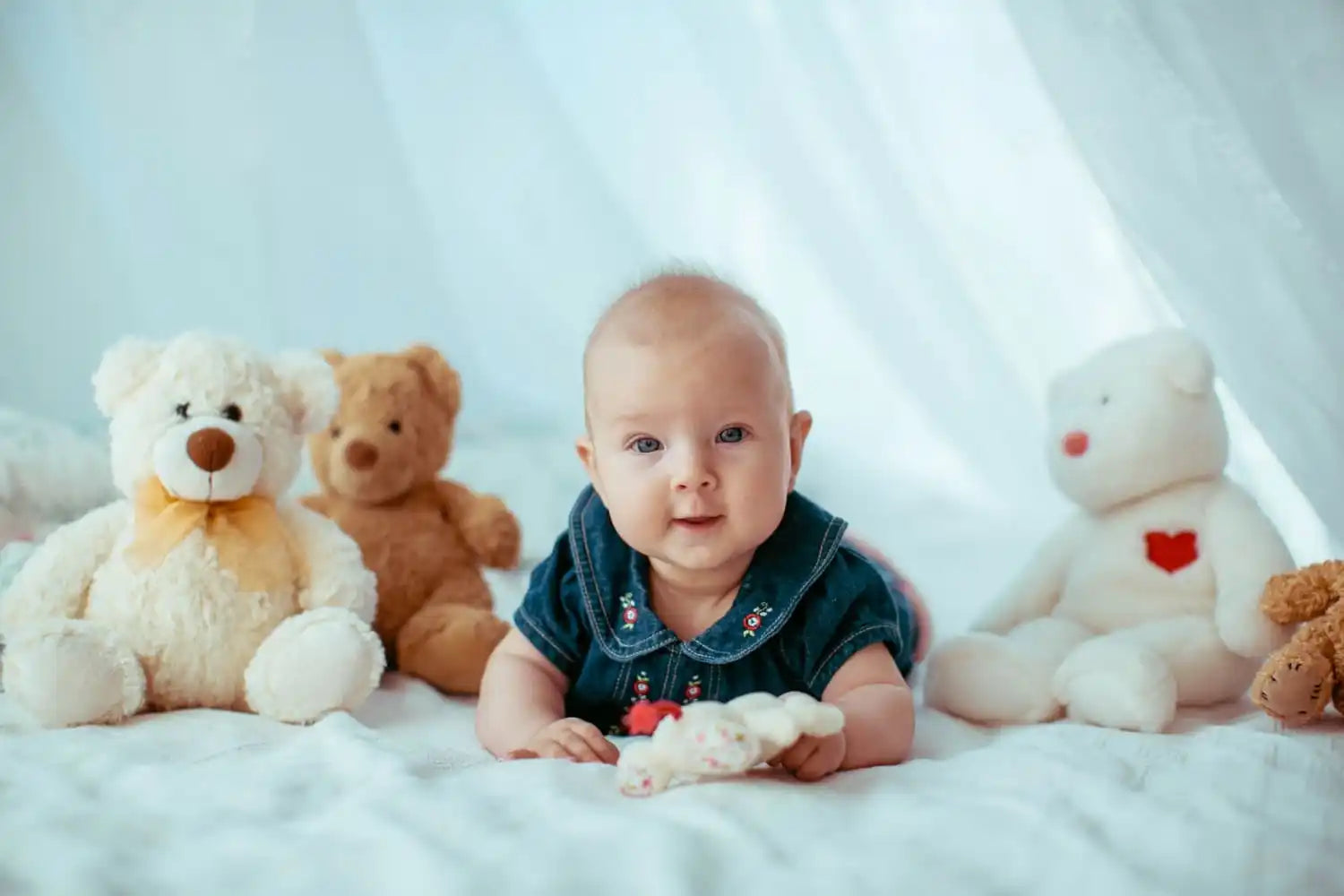 Un bébé en vêtements en denim allongé sur un tissu blanc entre des ours en peluche.